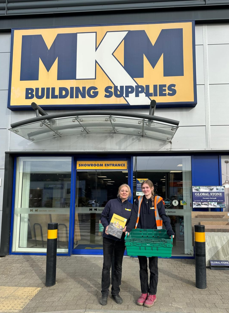 MKM Building Supplies Milton Keynes storefront with team receiving MK Food Bank donation crate outside entrance